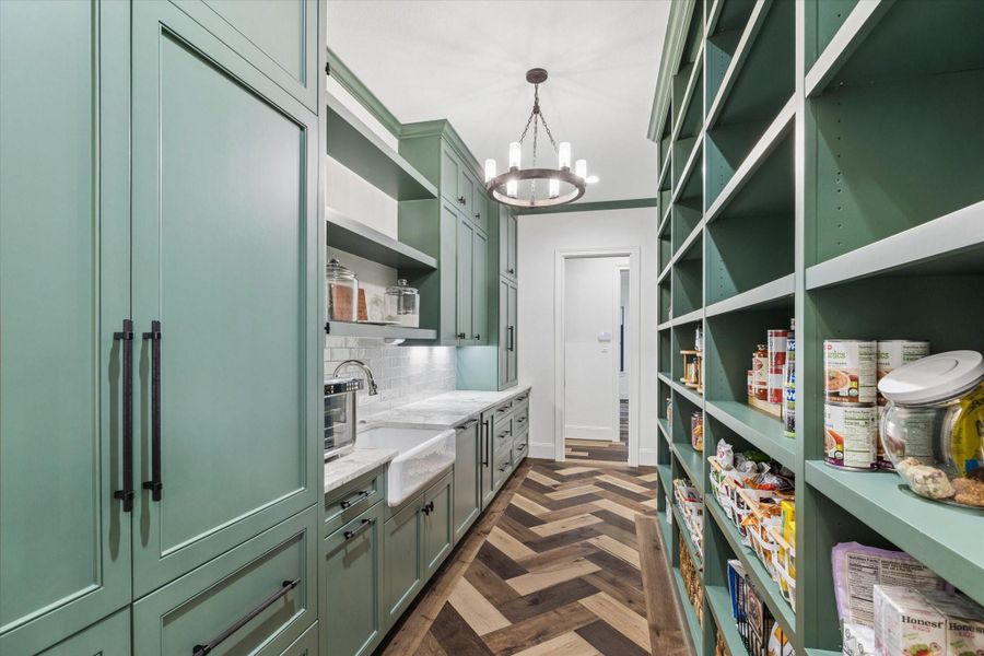 This additional view showcases the exceptional storage capacity and designer craftsmanship that define the prep pantry and scullery. Floor-to-ceiling cabinetry, expansive open shelving, pullout organization systems, and generous countertops ensure every item has its place. Rich green cabinetry, a handcrafted-inspired backsplash, farmhouse sink, designer lighting, and herringbone hardwood flooring create a sophisticated secondary kitchen that seamlessly complements the home’s luxurious main culinary space.