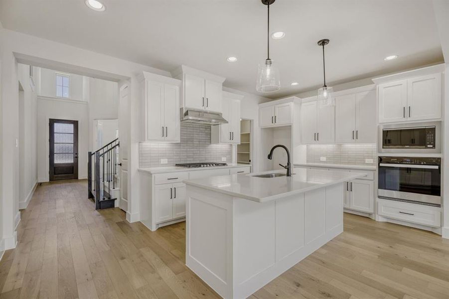 Kitchen with stainless steel appliances, light wood finished floors, white cabinetry, hanging light fixtures, and a center island with sink