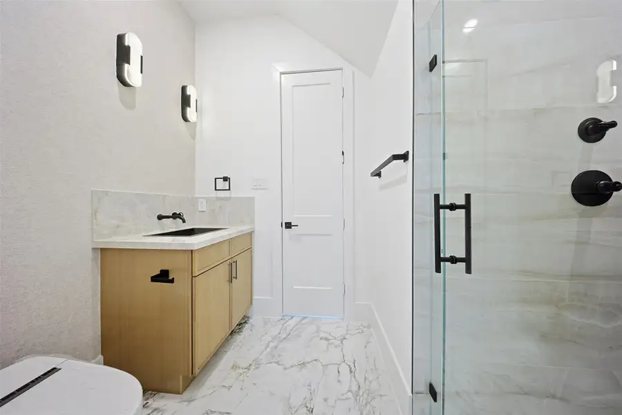 The bathroom offers a light wood vanity with a marble-patterned countertop, black fixtures, and contemporary wall sconces.