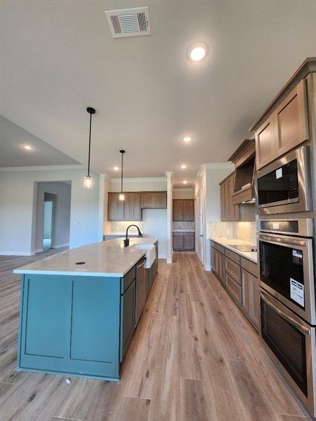 Open-concept kitchen featuring a large central island with a farmhouse sink, stainless steel appliances including double ovens, wood-finish flooring, recessed lighting, and pendant light fixtures