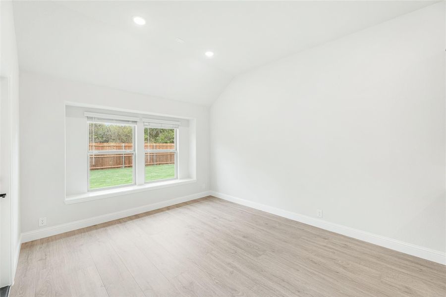 Empty bedroom with vaulted ceiling, light wood floors, and a window overlooking a fenced backyard