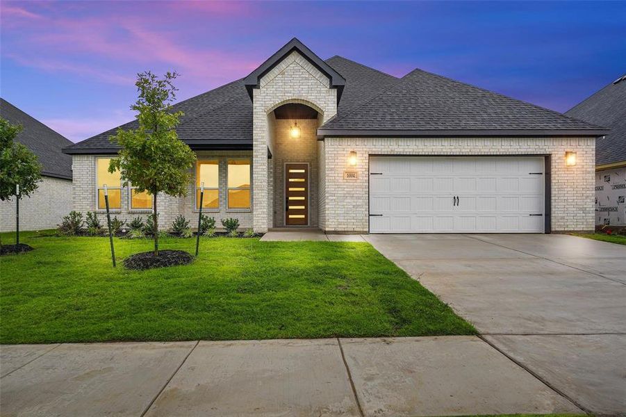 French country inspired facade featuring roof with shingles, brick siding, a garage, and driveway French country inspired facade featuring roof with shingles, brick siding, a garage, and driveway