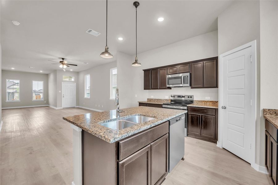 Kitchen with pendant lighting, a kitchen island with sink, dark wood finish cabinets, stainless steel appliances, and light stone countertops