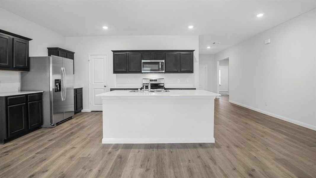 Kitchen featuring an island with sink, stainless steel appliances, light stone counters, dark cabinetry, and recessed lighting