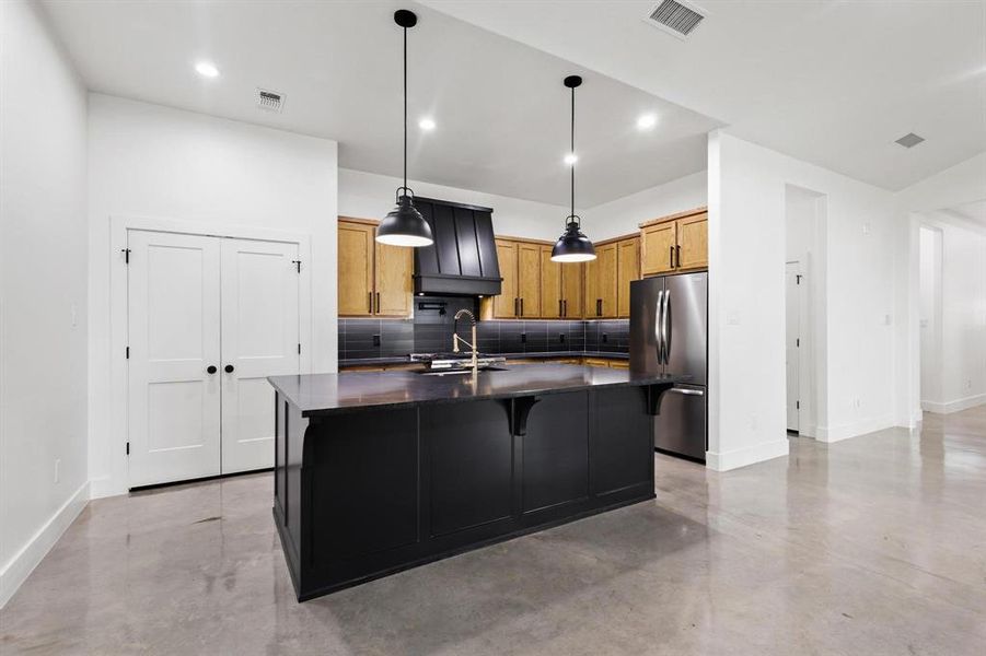 Kitchen featuring concrete flooring, tasteful backsplash, a kitchen island with sink, decorative light fixtures, and freestanding refrigerator