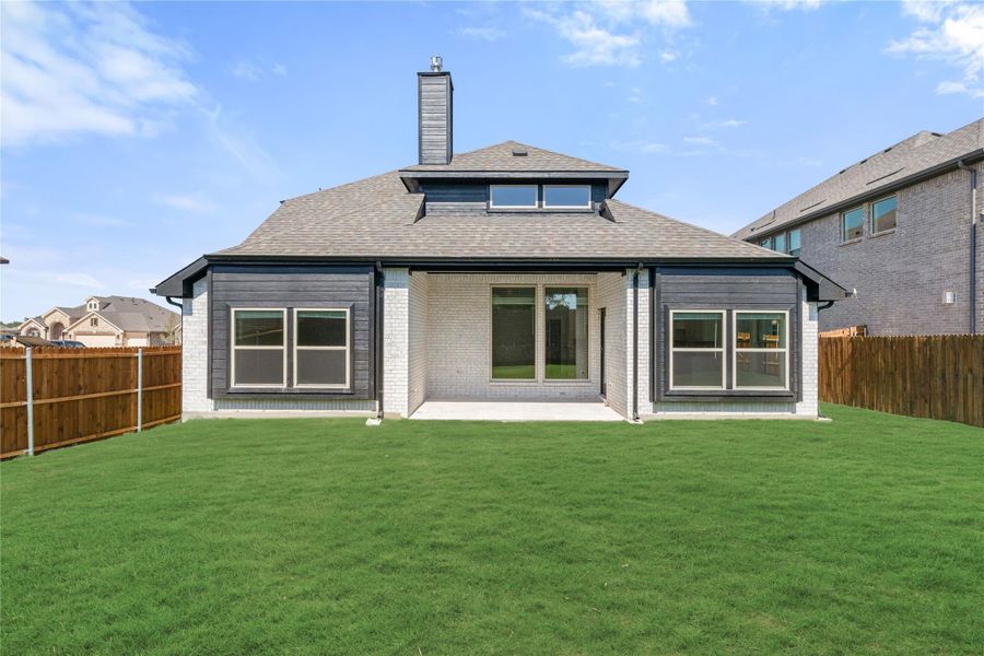 Rear exterior of single-story home with white brick, dark trim, covered patio, and fenced backyard with green lawn