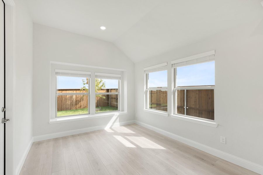 Empty bedroom with vaulted ceiling, light wood floors, and multiple windows overlooking fenced backyard