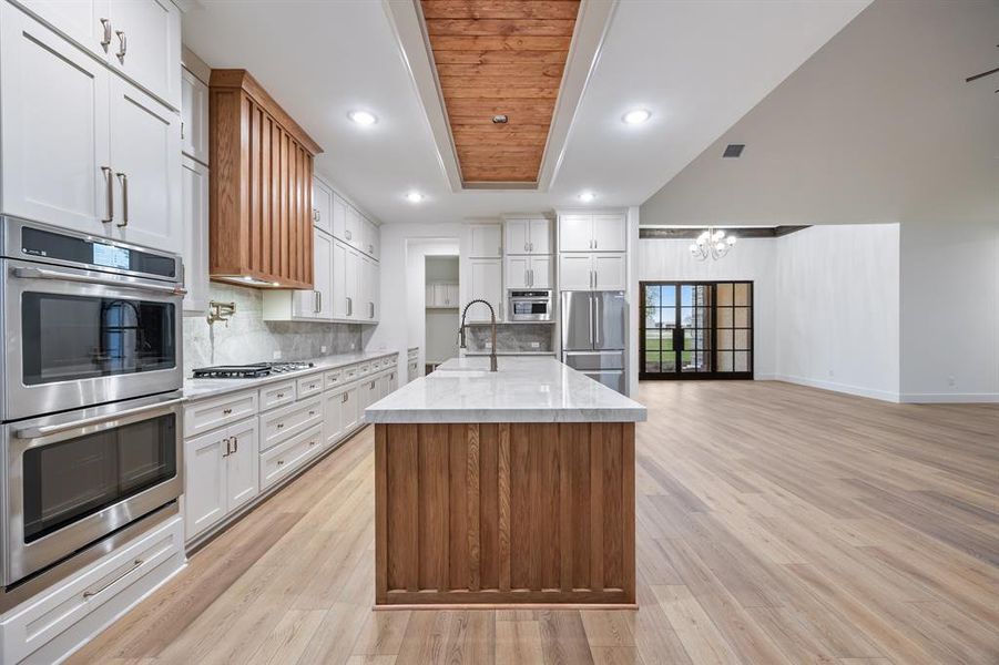 Kitchen featuring stainless steel appliances, white cabinetry, light stone counters, an island with sink, and brown cabinetry