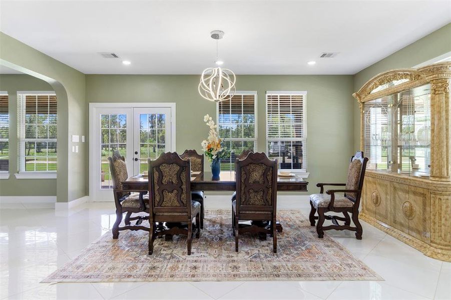 Bright, elegant dining room with large windows and French doors offering abundant natural light. Features a classic wooden dining set, ornate china cabinet, and modern light fixture. The space is accented with a stylish area rug and glossy tile flooring, creating a welcoming atmosphere.