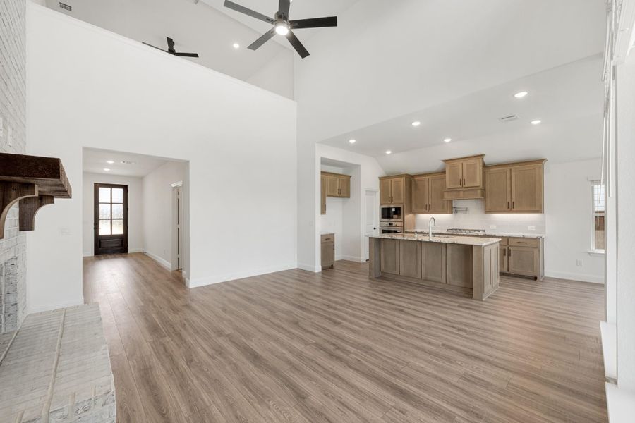 Open-concept living area with high ceilings, wood-look floors, kitchen island, tan cabinets, and brick fireplace Open-concept living area with high ceilings, wood-look floors, kitchen island, tan cabinets, and brick fireplace
