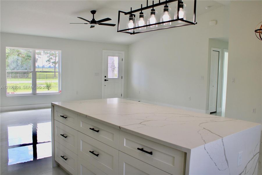 Large kitchen island featuring a veined stone countertop and white shaker-style cabinetry with matte black hardware, accented by a linear multi-light pendant fixture and a ceiling fan