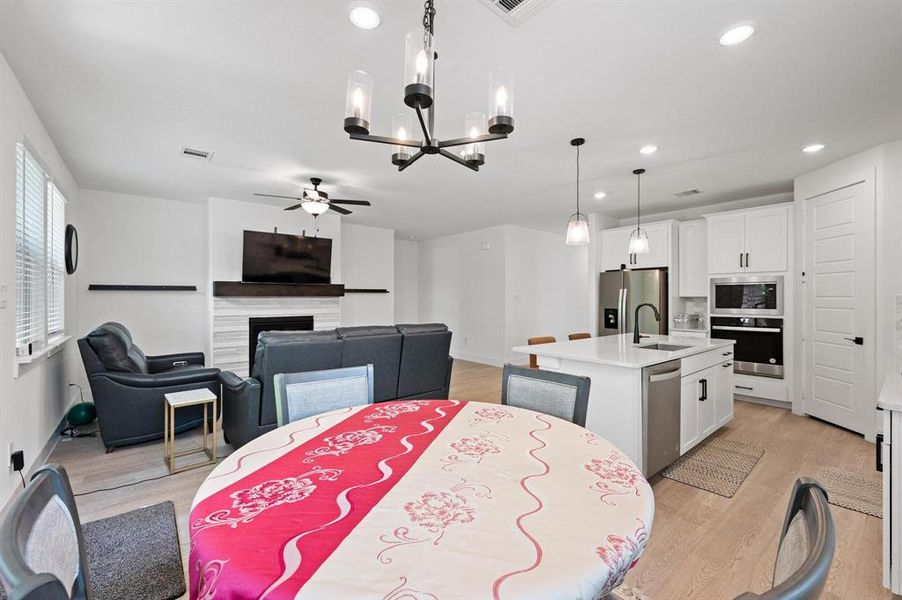 Open-concept interior featuring light wood-finish flooring, a white kitchen with stainless steel appliances, a central island with a sink, pendant lighting, and a living area with a fireplace Open-concept interior featuring light wood-finish flooring, a white kitchen with stainless steel appliances, a central island with a sink, pendant lighting, and a living area with a fireplace