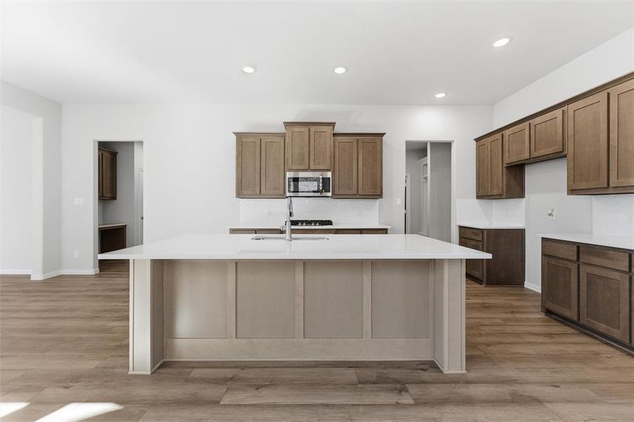 Kitchen featuring decorative backsplash, brown cabinetry, an island with sink, recessed lighting, and light stone countertops