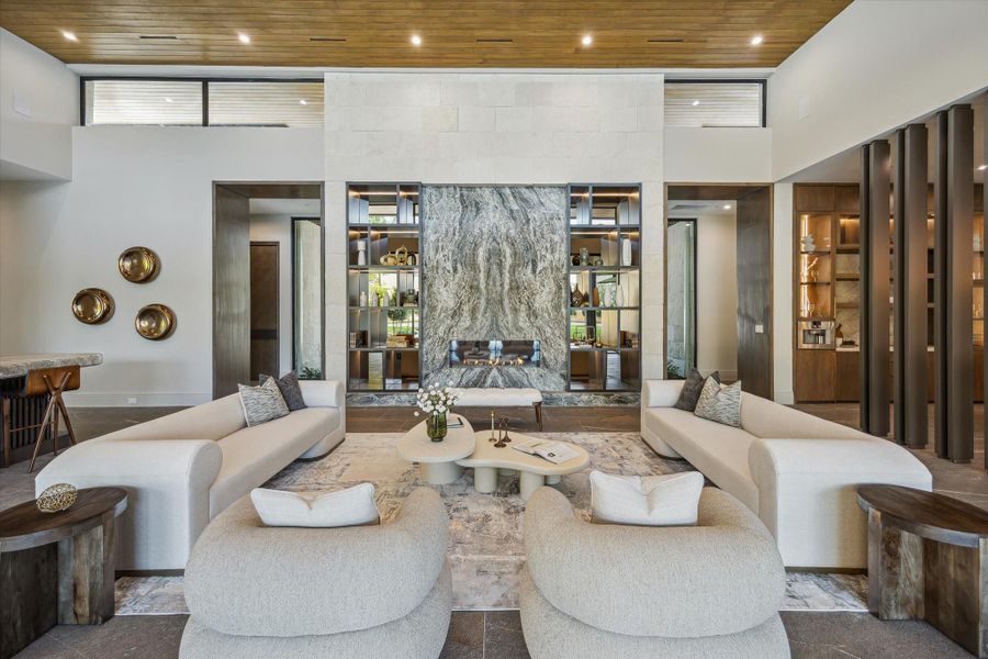 This exquisite living room is a true architectural showpiece, centered around a dramatic porcelain-surround fireplace with hand-selected Fantasy Brown Francini marble and custom built-ins. A soaring tray ceiling finished in warm wood planks, designer recessed lighting, and in-ceiling speakers complete the space with refined sophistication and style.