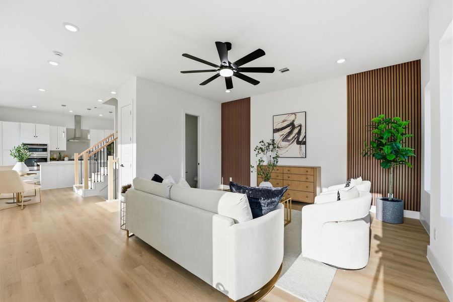 Open-concept living area featuring wood-look flooring, recessed lighting, a modern ceiling fan, and warm accent wood paneling, seamlessly flowing into the kitchen for a bright, stylish space ideal for everyday living and entertaining.
