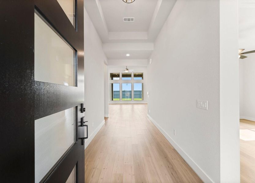 This photo showcases a modern entryway with light wood flooring leading to a bright, open living space. Large windows at the far end offer plenty of natural light and a view of the outdoors, creating a welcoming atmosphere.