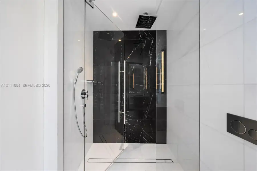Walk-in shower featuring glass enclosure, ceiling-mounted rainfall showerhead, handheld shower fixture, black stone-finish tile accent wall, and linear floor drain Walk-in shower featuring glass enclosure, ceiling-mounted rainfall showerhead, handheld shower fixture, black stone-finish tile accent wall, and linear floor drain