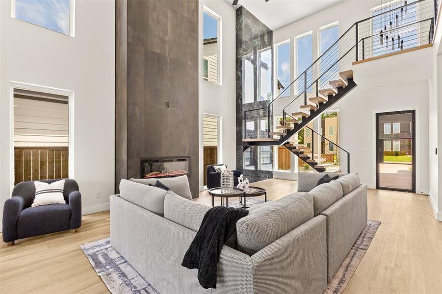 Two-story living area featuring a prominent fireplace wall, a floating staircase with cable railing, high ceilings, abundant natural light from multiple windows, and light wood-finish flooring