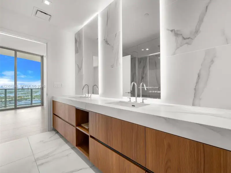Floating wood-finish vanity with dual integrated sinks, light-toned stone-finish countertop, full-height decorative stone-finish backsplash, and recessed LED mirror lighting Floating wood-finish vanity with dual integrated sinks, light-toned stone-finish countertop, full-height decorative stone-finish backsplash, and recessed LED mirror lighting