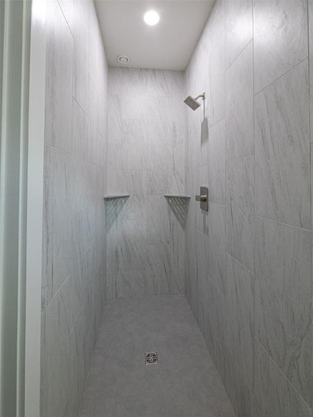 Walk-in shower featuring light grey veined tile walls, a matte grey tile floor, a brushed nickel shower head, and built-in shower niches
