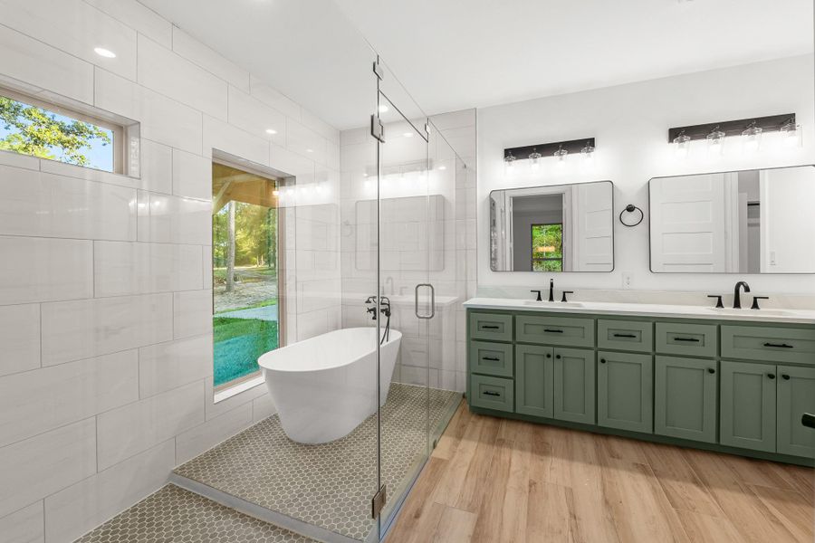 The primary bathroom features a modern design with a glass-enclosed shower and standalone tub, dual mirrors above a green double vanity, and elegant light fixtures. Large windows provide natural light and a view of the outdoors, complemented by stylish tile flooring. The primary bathroom features a modern design with a glass-enclosed shower and standalone tub, dual mirrors above a green double vanity, and elegant light fixtures. Large windows provide natural light and a view of the outdoors, complemented by stylish tile flooring.