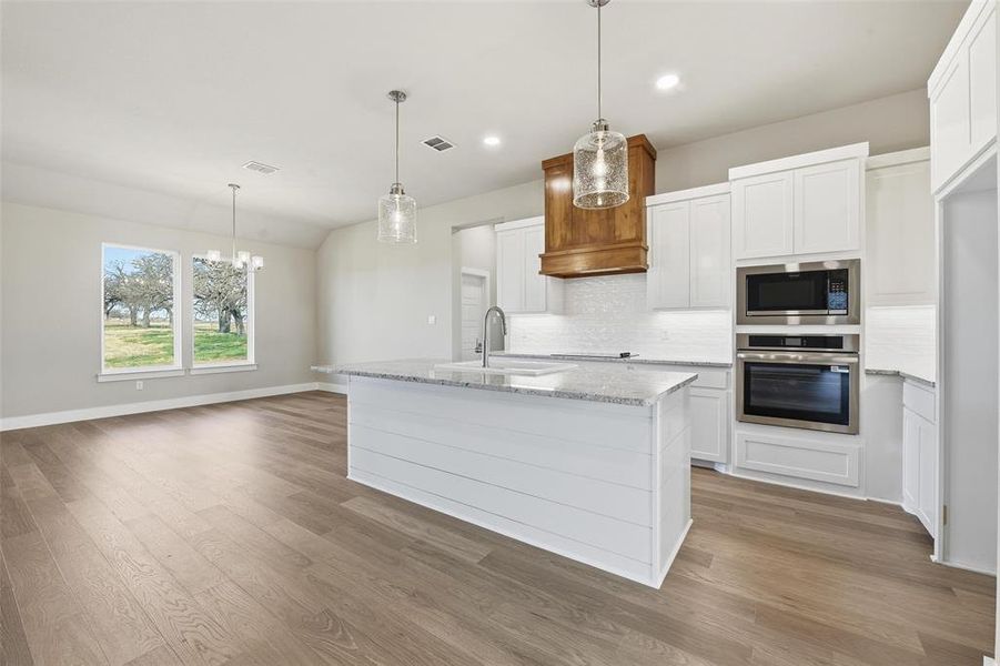 Kitchen featuring decorative light fixtures, white cabinetry, stainless steel appliances, light stone counters, and an island with sink