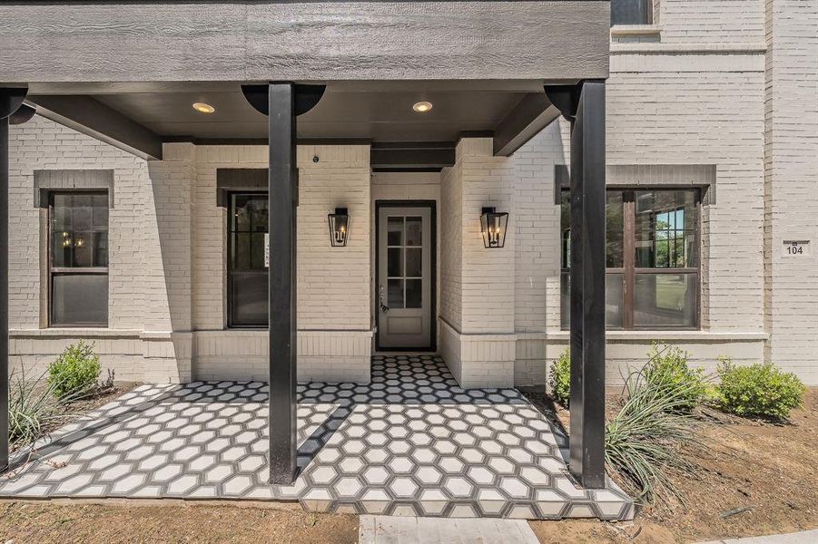 Entryway featuring a covered porch, white brick exterior, black hexagonal tile flooring, and two wall-mounted lanterns flanking the front door Entryway featuring a covered porch, white brick exterior, black hexagonal tile flooring, and two wall-mounted lanterns flanking the front door