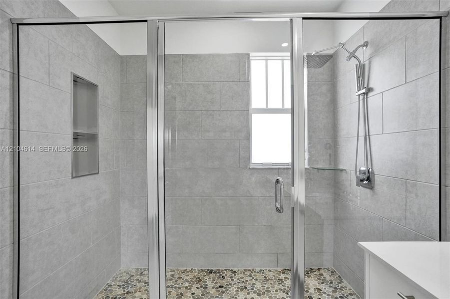 Shower with sliding glass enclosure, light gray tile walls, built-in recessed shelf, and pebble-finish flooring