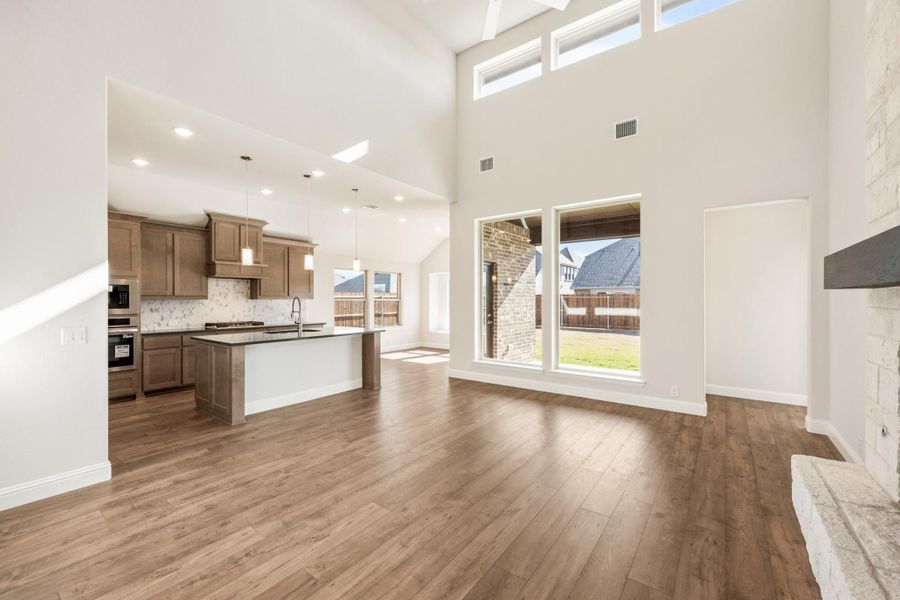 Open-concept living area with hardwood floors, vaulted ceilings, and kitchen with brown cabinets and island visible in background Open-concept living area with hardwood floors, vaulted ceilings, and kitchen with brown cabinets and island visible in background