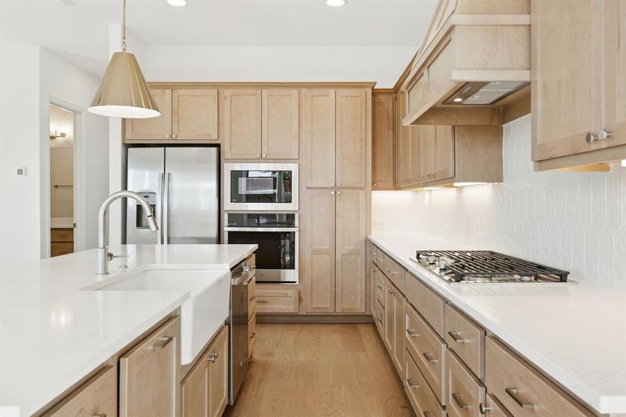 Kitchen featuring light brown cabinetry, appliances with stainless steel finishes, decorative light fixtures, light wood-type flooring, and light stone countertops