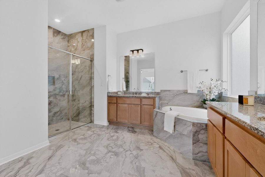 Spacious bathroom featuring large format stone-look tile flooring, an oversized walk-in shower with clear glass enclosure and matching stone-look tile, a built-in soaking tub with stone-look surround, and dual wood-finish vanities with granite countertops