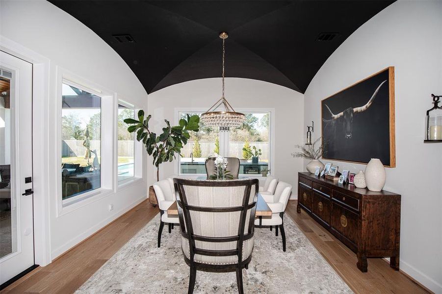 This dining area features a vaulted black ceiling and large windows that offer ample natural light. It includes a modern chandelier, a decorative sideboard, and a view of the backyard with a pool.