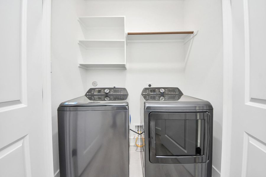 This photo shows a compact laundry room featuring modern, stainless steel washer and dryer units. There are open shelves and a hanging rod for convenience, with a clean, white aesthetic.