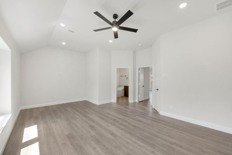 Empty master bedroom with vaulted ceiling, ceiling fan, wood floors, and ensuite bathroom doorway Empty master bedroom with vaulted ceiling, ceiling fan, wood floors, and ensuite bathroom doorway