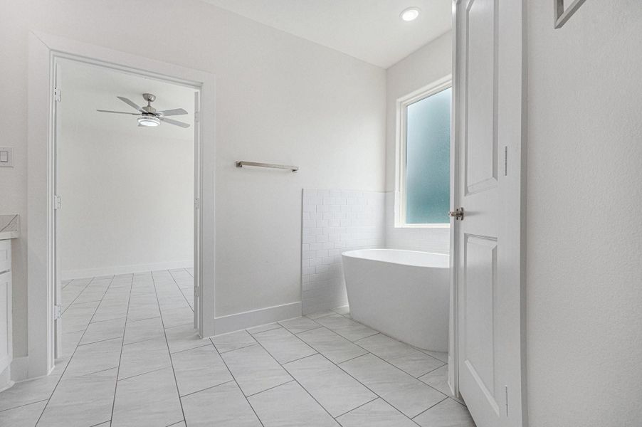This photo showcases a bright, modern bathroom with a standalone bathtub, large frosted window for privacy, and sleek tiled flooring. The space connects to an adjoining room with a ceiling fan, offering an open and airy feel. This photo showcases a bright, modern bathroom with a standalone bathtub, large frosted window for privacy, and sleek tiled flooring. The space connects to an adjoining room with a ceiling fan, offering an open and airy feel.