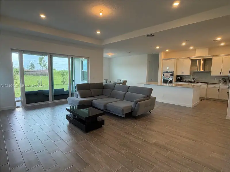 Open-concept space featuring wood-finish tile flooring, recessed ceiling lighting, a tray ceiling, sliding glass doors, and kitchen cabinetry with a built-in cooktop range