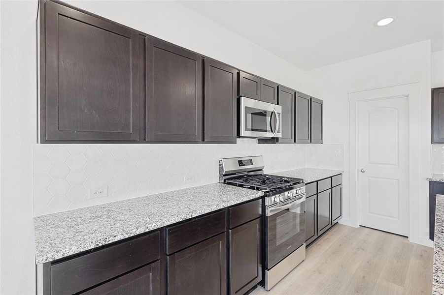 Kitchen with appliances with stainless steel finishes, tasteful backsplash, dark brown cabinets, light hardwood / wood-style floors, and light stone counters