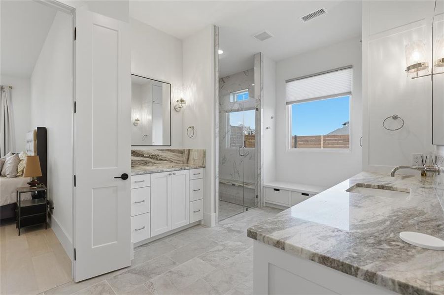 Bathroom featuring dual white vanities with stone countertops, a glass-enclosed shower with bench seating, a window with a privacy blind, and large format tile flooring