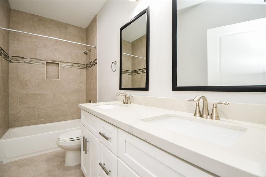 This is a modern, clean-lined secondary bathroom featuring a double vanity with undermount sinks, sleek brushed nickel faucets, a large mirror, a bathtub/shower combo with tile surround, and a toilet. The neutral color palette provides a calming atmosphere.