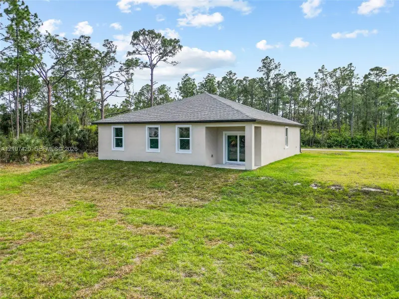 Rear elevation featuring a stucco exterior, hipped roof, sliding glass doors, and expansive grassy grounds surrounded by mature trees Rear elevation featuring a stucco exterior, hipped roof, sliding glass doors, and expansive grassy grounds surrounded by mature trees