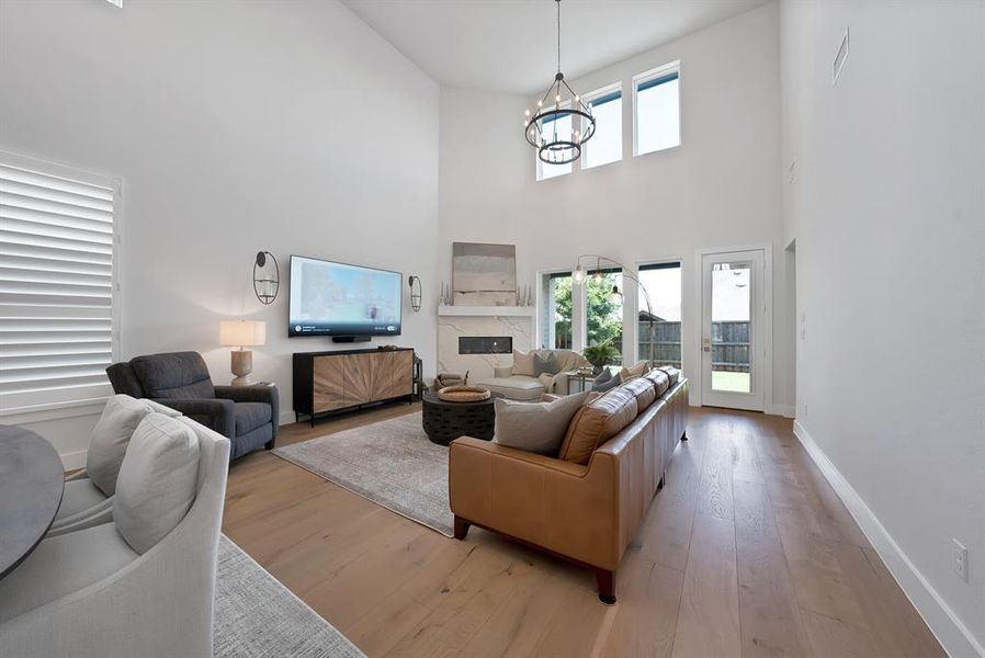 Spacious living area featuring high vaulted ceilings, large windows, light wood-finish flooring, a contemporary chandelier, and a built-in fireplace with a mantel Spacious living area featuring high vaulted ceilings, large windows, light wood-finish flooring, a contemporary chandelier, and a built-in fireplace with a mantel