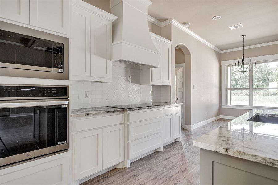 Kitchen with stainless steel appliances, white cabinetry, light stone counters, premium range hood, and a chandelier