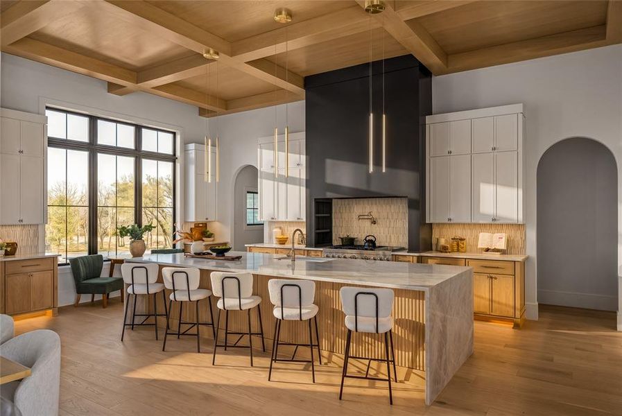 Gourmet kitchen featuring a large central island with waterfall countertop, coffered ceiling, expansive black-framed windows, integrated range with custom hood, and light wood-finish flooring
