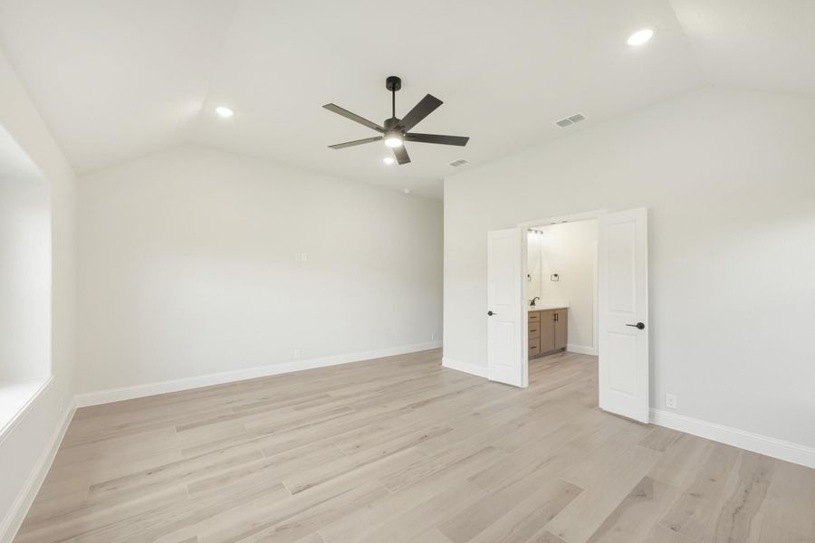 Empty bedroom with vaulted ceiling, ceiling fan, light wood floors, and ensuite bathroom doorway visible.