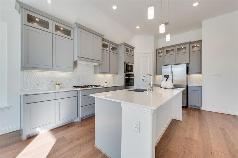 Kitchen with light wood-type flooring, stainless steel appliances, sink, decorative light fixtures, and an island with sink