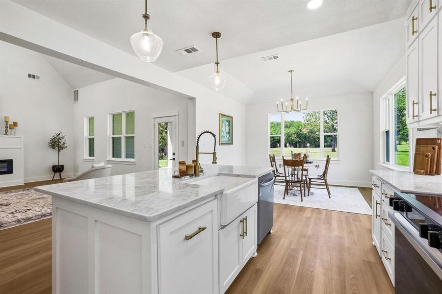 Open-concept interior featuring a kitchen island with stone countertop and farmhouse sink, wood-finish flooring, white cabinetry with gold-tone hardware, and a dining area with expansive window Open-concept interior featuring a kitchen island with stone countertop and farmhouse sink, wood-finish flooring, white cabinetry with gold-tone hardware, and a dining area with expansive window