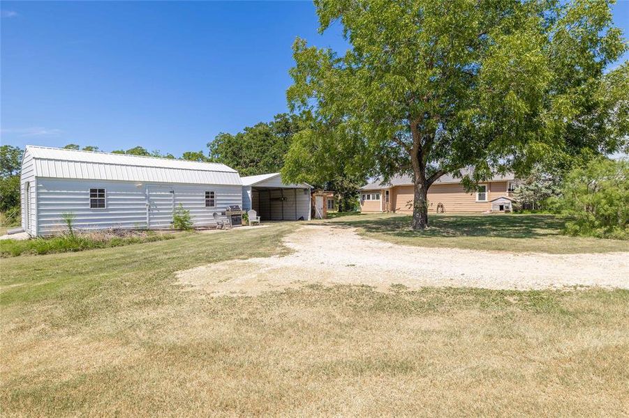 View of green lawn with a carport, dirt driveway, and an outdoor structure View of green lawn with a carport, dirt driveway, and an outdoor structure