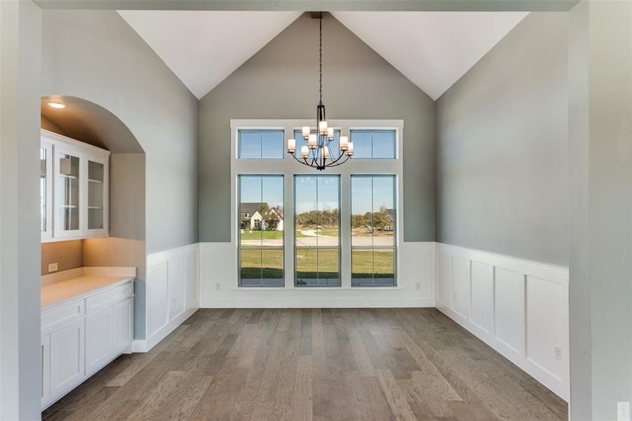 Unfurnished dining area featuring light wood finished floors, high vaulted ceiling, a chandelier, and a wainscoted wall Unfurnished dining area featuring light wood finished floors, high vaulted ceiling, a chandelier, and a wainscoted wall