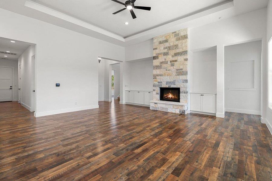 Unfurnished living room with ceiling fan, a tray ceiling, a stone fireplace, dark wood-style floors, and recessed lighting