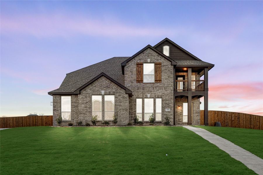 Two-story brick home with second-floor balcony, large windows, and green lawn at dusk Two-story brick home with second-floor balcony, large windows, and green lawn at dusk
